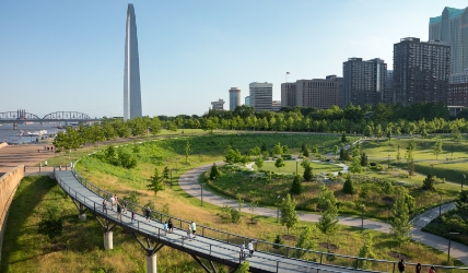 Image of North Gateway area of Gateway Arch National Park. It includes a view of the St. Louis skyline from the northern area of the city, the Explorer's Garden and paved pathways with bikers, walkers and runners on them.